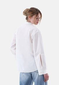 Camicia bianca a maniche lunghe con colletto tradizionale, aperture laterali e polsini abbottonati, abbinata a jeans di denim azzurro chiaro.
