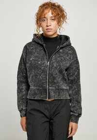 Sudadera recortada negra de tejido suave y texturizado con cierre de cremallera, capucha con cordón y detalles de costura en contraste. Combinada con pantalones negros.