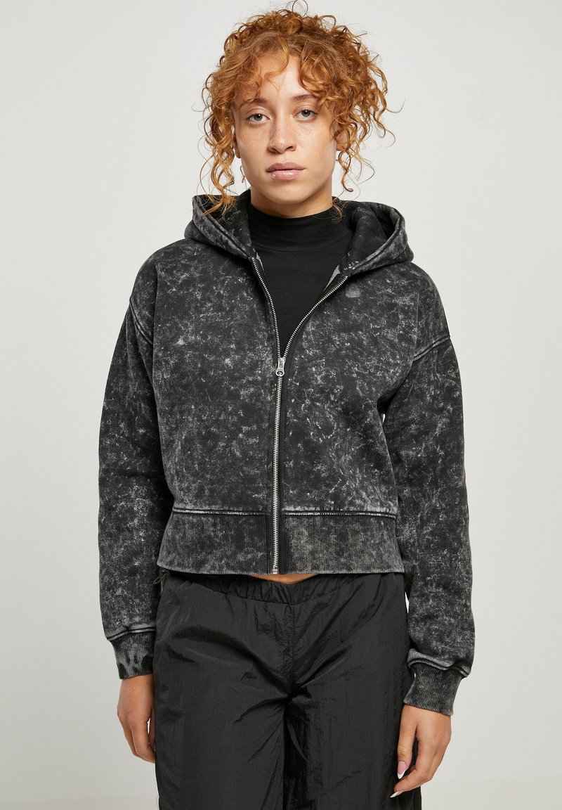 Sudadera recortada negra de tejido suave y texturizado con cierre de cremallera, capucha con cordón y detalles de costura en contraste. Combinada con pantalones negros.
