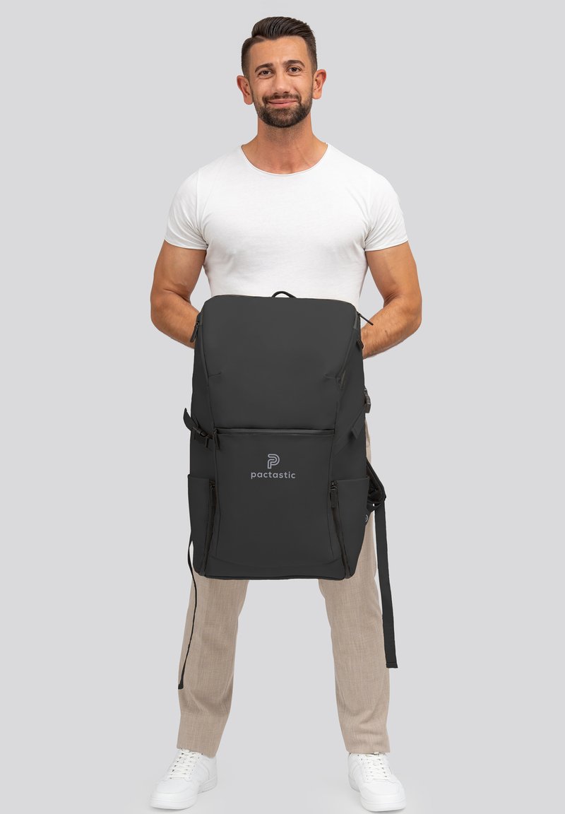 Sac à dos noir avec plusieurs poches zippées, texture lisse, bretelles réglables et un logo sur le devant. Conception robuste, spacieux.