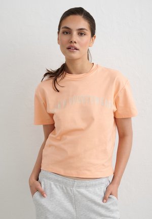 Ung kvinde med brunt hår i en hestehale iført en ferskenfarvet T-shirt med korte ærmer og lysegrå joggingbukser, står foran en ensfarvet hvid væg.