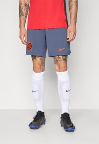 T-shirt de sport rouge, shorts bleus avec le logo Nike et l'emblème de l'équipe, chaussettes blanches montantes avec des détails violets, chaussures de football noires avec des accents bleus.