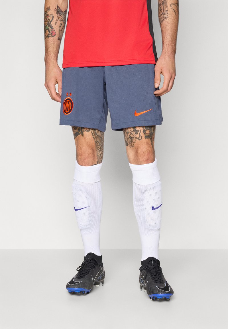 Rød idrettsskjorte, blå shorts med Nike-logo og lagemblemet, hvite knelange sokker med lilla detaljer, svarte fotballsko med blå aksenter.