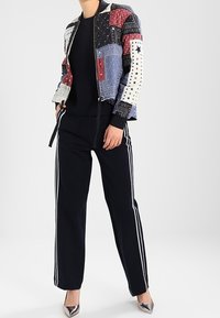 Veste bomber patchwork en rouge, bleu et blanc avec des motifs variés ; associée à un pantalon noir avec des rayures blanches sur les côtés ; talons argentés.