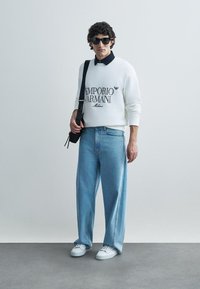 Sweatshirt branco com logótipo preto "Emporio Armani", camisa azul-marinho com gola, jeans claros e largos, ténis brancos e óculos de sol pretos.