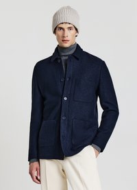 Giacca blu navy in misto lana con chiusura con bottoni, due tasche frontali e colletto, abbinata a un berretto a coste grigio. Pantaloni chiari di colore neutro.