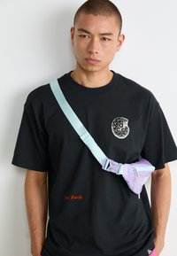 Mand i sort T-shirt med fossilgrafik og orange tekst, accessoriseret med lys blå rem og en lille lilla crossover-taske.