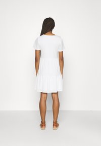 Bílá bavlněná šaty s krátkými rukávy a vrstevnatou sukní, s kulatým výstřihem a hladkou texturou. Nosené s hnědými sandály.