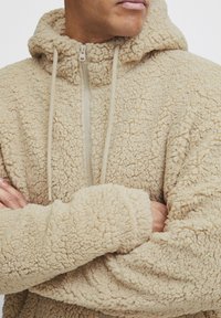 Beige fleecejakke med en teksturert, fluffy finish, hel glidelås og hette. Har snor og løstsittende ermer.