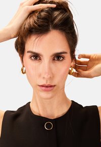 Orecchini a cerchio in oro, top senza maniche nero con un dettaglio in metallo circolare, pelle liscia, capelli castani raccolti, sfondo neutro.