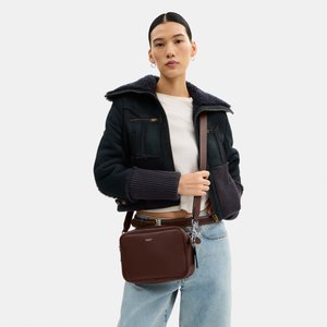 Žena u crnoj jakni od ovčjeg krzna i svijetlim trapericama, drži smeđu kožnu torbu preko ramena s logom sprijeda.