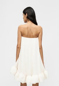 Γυναίκα με μακριά σκούρα μαλλιά φοράει λευκό, αμάνικο, φαρδύ φόρεμα με βολάν στο τελείωμα, στέκεται με την πλάτη στραμμένη στην κάμερα σε απλό φόντο.