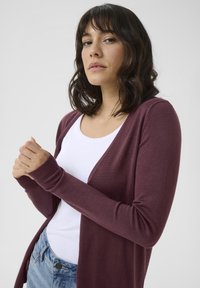 Burgunderfarget cardigan med lange ermer, åpen frontdesign, lett tekstur, båret over en hvit topp, som fremhever feminine konturer.