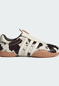Cow-print slip-on shoe featuring a smooth white leather upper, three white stripes, and a tan rubber sole. Rounded toe design.