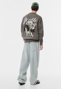 Barna pulóver, amelyen a "Death Valley" felirat látható fehér és zöld színben, világoskék laza farmernadrággal és fekete cipővel párosítva.