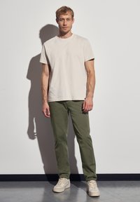 Vaaleanbeige lyhythihainen t-paita, joka on yhdistetty oliivinvihreisiin slim-fit-housuihin ja kermaisiin lenkkareihin valkoista seinää vasten.