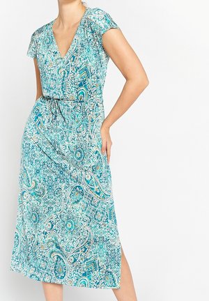 Kobieta ubrana w turkusowo-białą sukienkę midi w krótkim rękawie z wzorem, dekoltem w serek i wiązaniem w talii, stojąca z jedną ręką na biodrze.