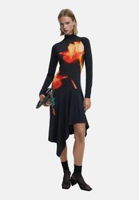 Langärmliges Kleid mit einer taillierten Oberseite und asymmetrischem Saum, das ein florales orangefarbenes Muster auf dunklem Hintergrund zeigt. Begleitet von einer strukturierten Handtasche.