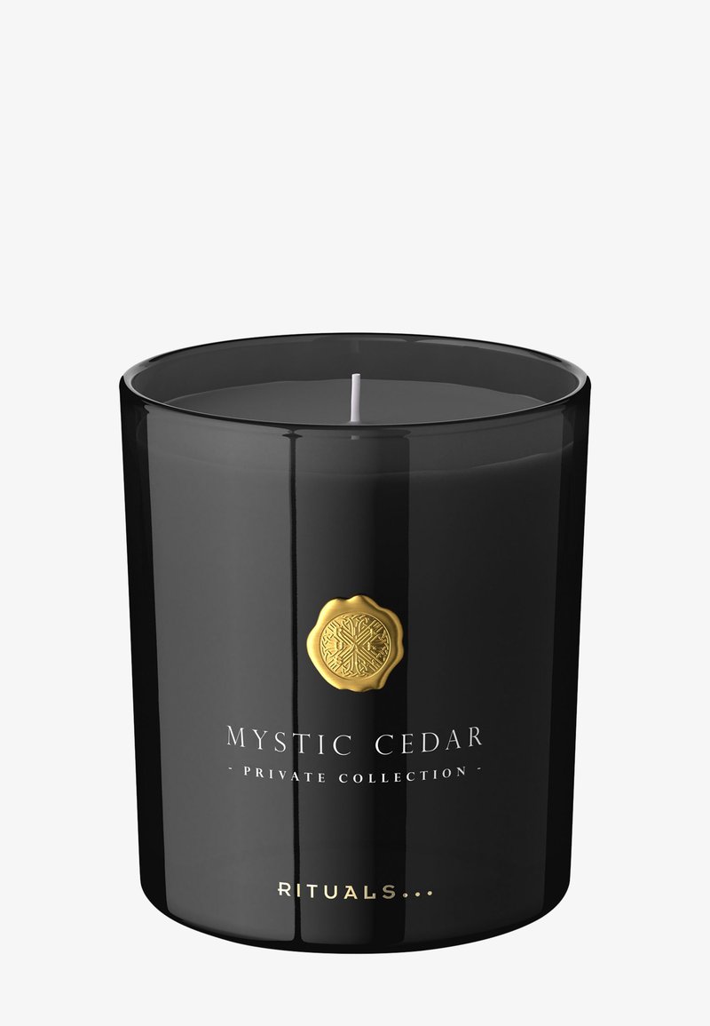Musta lasinen kynttilänjalka, jossa on kultainen vahalabeli, jossa lukee "MYSTIC CEDAR". Valkoinen kynttilä sisällä, sileä pinta, pyöreä muoto.
