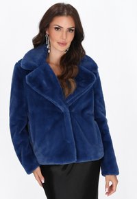Kvinde med langt bølget hår iført store perle- og ædelstensøreringe, en blød blå pelsjakke i fake fur og en sort nederdel, der poserer mod en hvid baggrund.