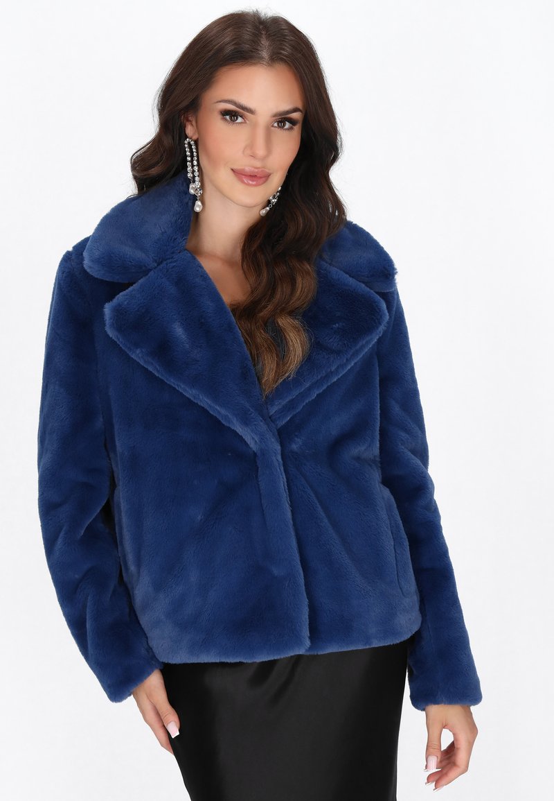 Kvinde med langt bølget hår iført store perle- og ædelstensøreringe, en blød blå pelsjakke i fake fur og en sort nederdel, der poserer mod en hvid baggrund.
