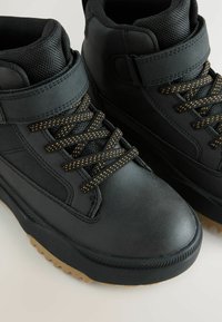 Svarta ankle boots med texturerat läder, diamantmönstrade snören med gulddetaljer och gummisula. Har en vadderad krage.