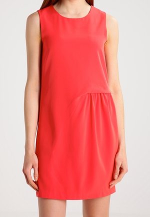Mouwloze koraalrode shiftjurk met ronde hals en geplooid detail aan de rechterzijde onder de taille, gedragen door een staand persoon.