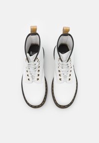 Botas de tobillo de cuero blanco con detalles en negro, costuras amarillas, puntera redonda y ojales dorados. Presentan una lengüeta en la parte posterior.