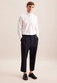 Camicia bianca aderente con colletto e taschino. Pantaloni blu navy con pieghe e gamba affusolata. Scarpe in pelle nere. Sfondo leggero e neutro.