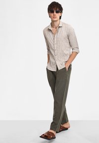 Camicia tessuta di un beige chiaro con maniche arrotolate, abbinata a pantaloni larghi di un verde oliva e sandali marroni senza chiusura. Gli occhiali da sole completano il look.
