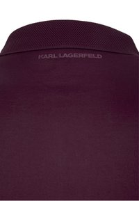 Burgundy-paita, jossa on tekstiiliä jäljittelevä kaulus. Takana on kohotettu logo "KARL LAGERFELD". Tasainen kangas peittää paidan. 