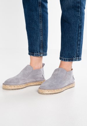 Person iført cropped blå jeans og lysegrå ruskind slip-on sko med vævede jutefødder, der står på et hvidt gulv.