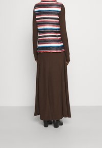 Ruskea pitkähihaista mekkoa on yhdistetty moniväriseen raidalliseen toppaliiviin. Liivissä on vaakasuoria raitoja sinisessä, vaaleanpunaisessa ja valkoisessa. Mustat jalkineet.