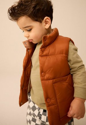 Gilet imbottito marrone con colletto alto, bottoni a pressione, indossato sopra una camicia a maniche lunghe verde chiaro e pantaloni a quadretti bianco e nero.