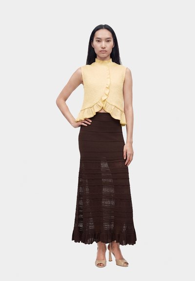 Femme aux longs cheveux noirs portant un top jaune sans manches à volants et une longue jupe marron foncé semi-transparente, debout avec une main sur la hanche.