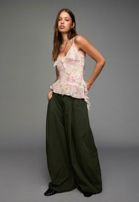 Floral-patterned, sheer, white camisole with ruffles paired with oversized, olive green trousers. Model poses with hands in pockets.