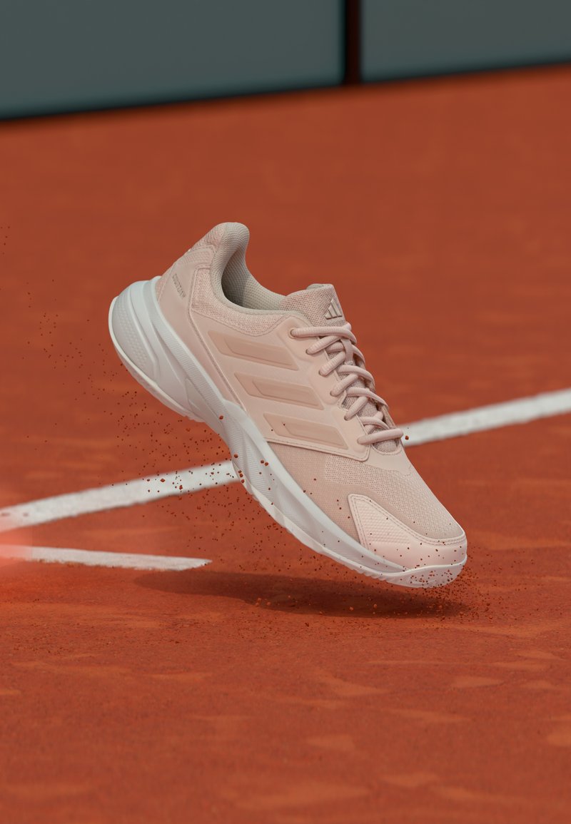 Scarpa da ginnastica rosa chiaro con tomaia testurizzata, intersuola bianca e suola in gomma con motivo, mostrata a mezz'aria su una superficie di terra battuta.