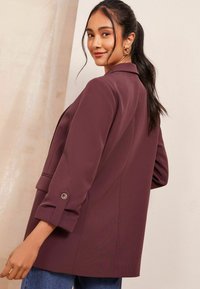 Fialový blazer s přizpůsobeným střihem, klopami, ohrnutými rukávy, knoflíkovými akcenty a hladkou texturou látky. Stylizováno s modrými džíny.