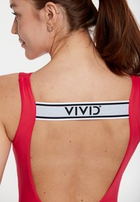 Červené jednodielne plavky s nízkym zadným dielom a bielym pásom s názvom značky "VIVID" v čiernej farbe. Hladký, štíhly materiál s priliehavým strihom.