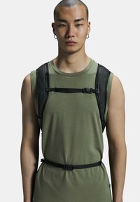 Black harness over an olive green sleeveless top, featuring straps, buckles, and a smooth texture. The harness is designed for a fitted look.