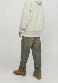 Persona che indossa una felpa leggera beige, jeans verdi sbiaditi e larghi, e stivaletti da lavoro color sabbia, in piedi con la schiena rivolta verso uno sfondo semplice.
