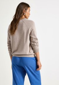 Beiger Pullover mit weicher Textur und gerippten Bündchen, kombiniert mit blauen Hosen. Der Pullover hat einen runden Ausschnitt und eine taillierte Passform.