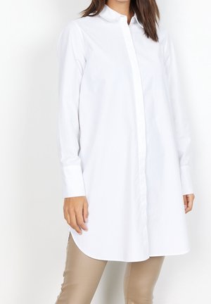 Femme portant une chemise blanche à manches longues boutonnée et un pantalon beige, debout devant un fond blanc.