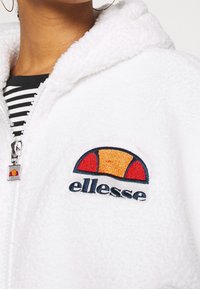 White fleece hoodie with a front zipper. Features an embroidered multicolored logo on the chest and a soft, textured finish. Striped shirt underneath.