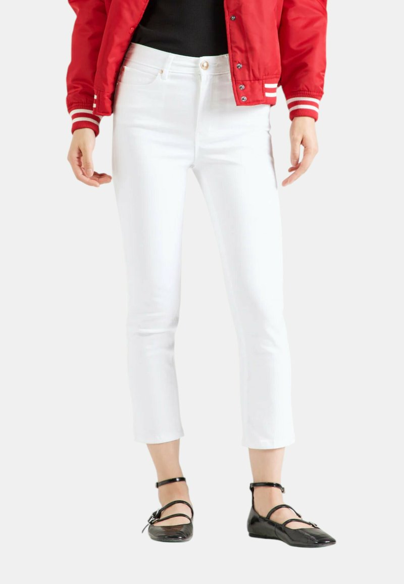 Jeans blancs coupés avec une coupe slim, présentant des coutures visibles et des poches à l'avant, associés à des ballerines en cuir verni noir avec des brides autour de la cheville.