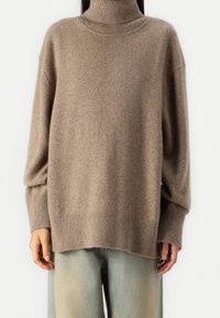 Maglione beige con collo alto realizzato in morbido tessuto a maglia, caratterizzato da una vestibilità ampia, polsini e orlo a costine. Abbinato a jeans a gamba larga di un blu chiaro.