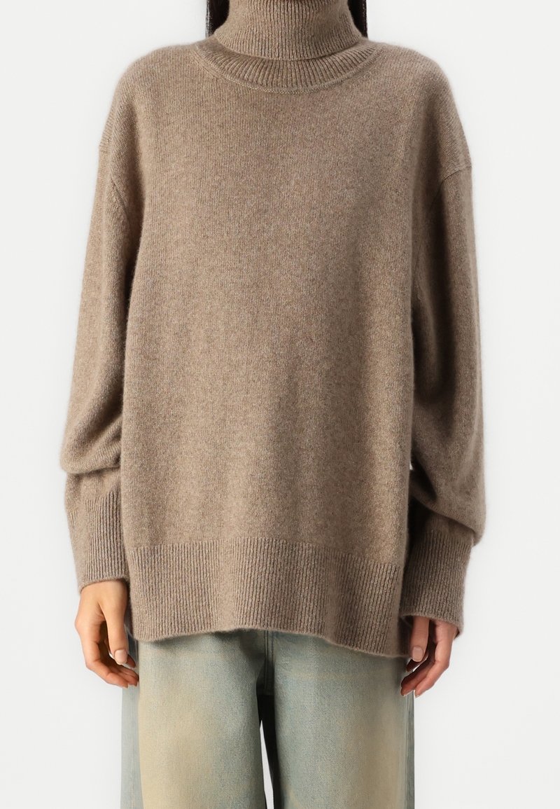 Maglione beige con collo alto realizzato in morbido tessuto a maglia, caratterizzato da una vestibilità ampia, polsini e orlo a costine. Abbinato a jeans a gamba larga di un blu chiaro.