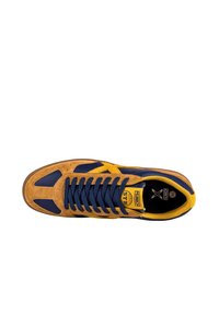 Zapatilla con upper de ante marrón claro, acentos en azul marino y detalles en amarillo. Presenta un diseño de cordones planos y una puntera redondeada.