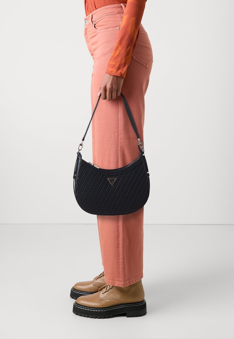 Borsa a tracolla lavorata in navy con una forma arrotondata e logo sul frontale. Abbinata a pantaloni a vita alta rosa chiaro e scarpe beige con lacci.