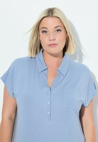 Camicia collare blu chiaro con maniche corte, botoniera frontale e una piccola tasca sul petto; realizzata in un tessuto liscio e leggero.